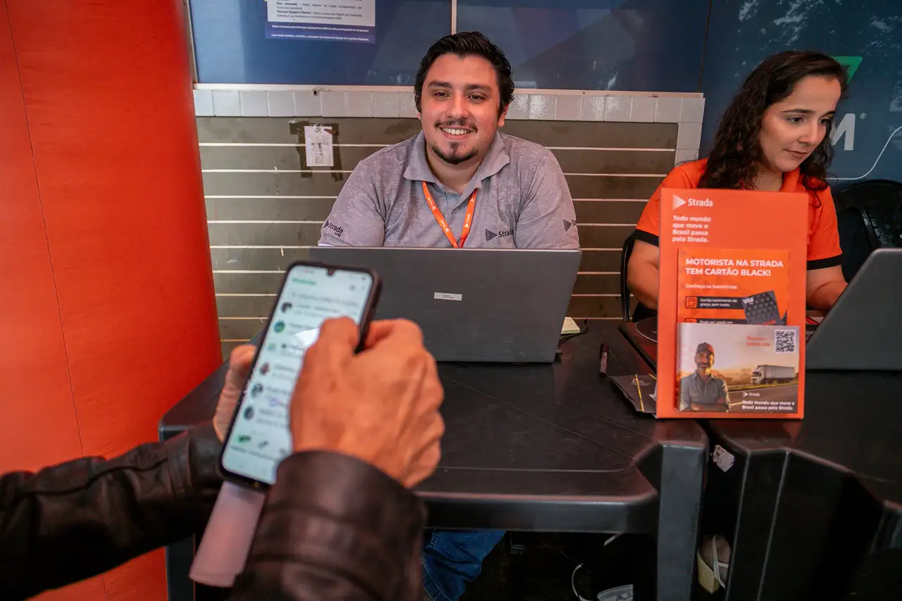Dois atendentes da Strada sorrindo no balcão de atendimento, com a mão de um caminhoneiro segurando o celular em primeiro plano.